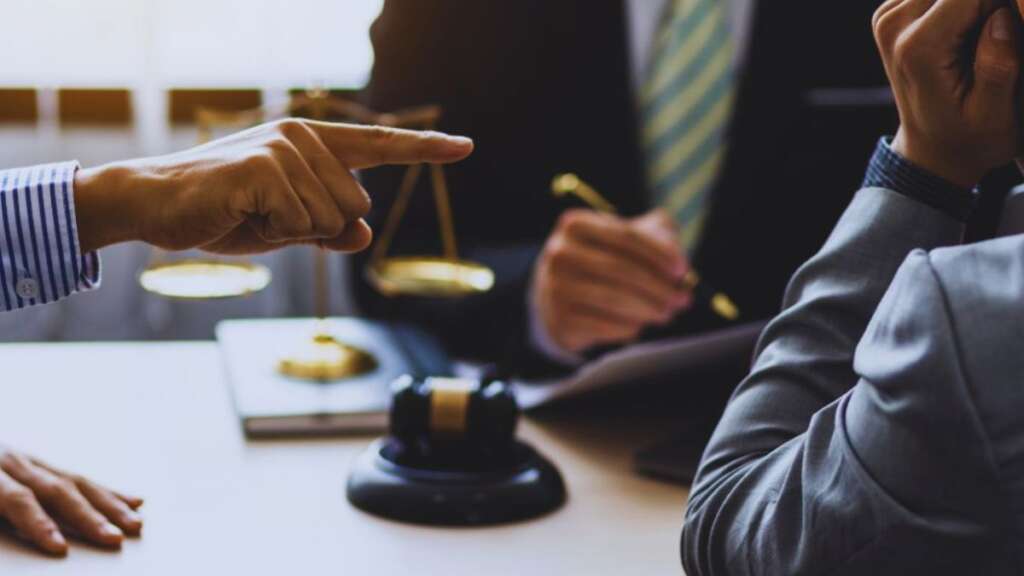 A gavel is placed between two model houses on a wooden table, representing a legal dispute over property ownership or title disagreements. A person in a blue shirt is seated in the background.