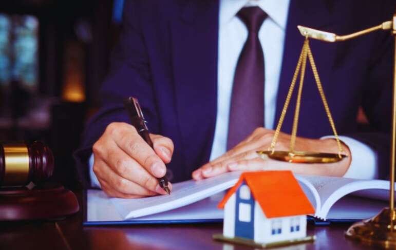 A property dispute litigation lawyer in a suit writing in a legal document, with a gavel, scales of justice, and a small model house on the desk, symbolizing real estate legal services.