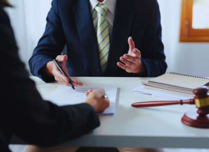 A contract lawyer in a business suit advises a client at a desk, reviewing a legal contract with a gavel and notebooks visible during a legal consultation.