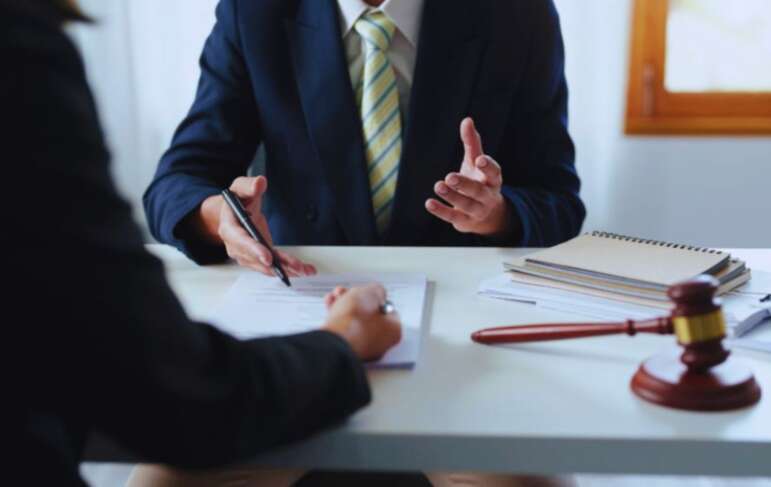 A contract lawyer in a business suit advises a client at a desk, reviewing a legal contract with a gavel and notebooks visible during a legal consultation.