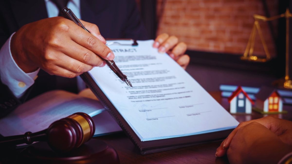 A contract lawyer in a suit is reviewing a real estate contract with a client across a desk, pointing to a section for signature. The scene includes a gavel, tiny model houses, and legal scales, emphasizing the lawyer’s role in real estate transactions and legal guidance.