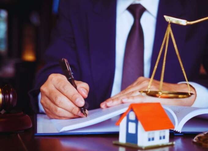 A professional estate planning lawyer in a suit sits at a desk, writing in a document with a pen. The desk features a judge’s gavel, scales of justice, and a small model house, symbolizing the role of an estate planning lawyer handling legal matters related to property and inheritance.
