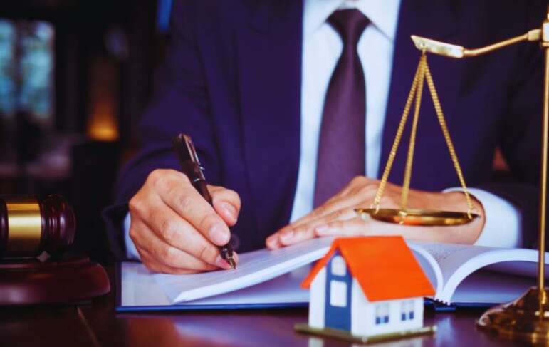 A professional estate planning lawyer in a suit sits at a desk, writing in a document with a pen. The desk features a judge’s gavel, scales of justice, and a small model house, symbolizing the role of an estate planning lawyer handling legal matters related to property and inheritance.