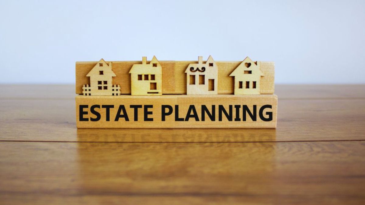 A wooden block with the words "ESTATE PLANNING" printed in black sits on a table. Four small wooden house figures are displayed on top of the block, representing different styles of homes against a plain, light-colored background.