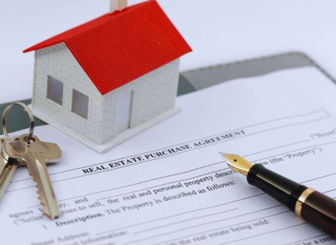 A real estate purchase agreement sits on a desk with a fountain pen, keys, and a model house, symbolizing the home-buying process.
