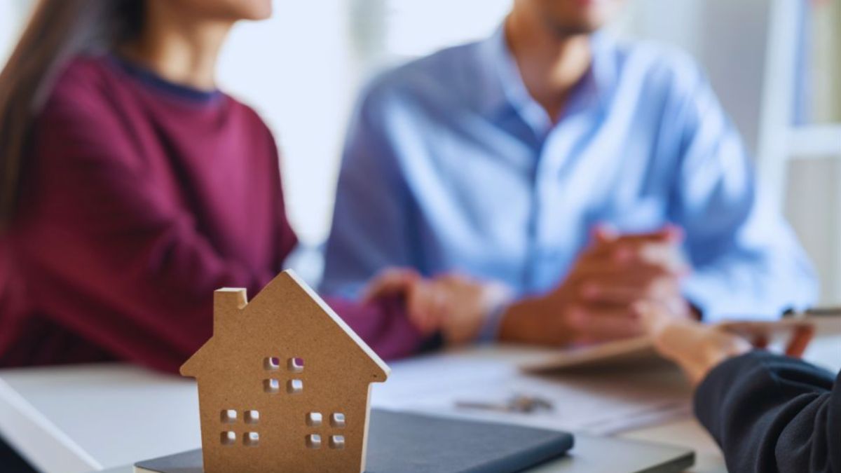 A small wooden house model sits on a table with a couple and a real estate agent in the background discussing documents and pens, illustrating a real estate transaction.