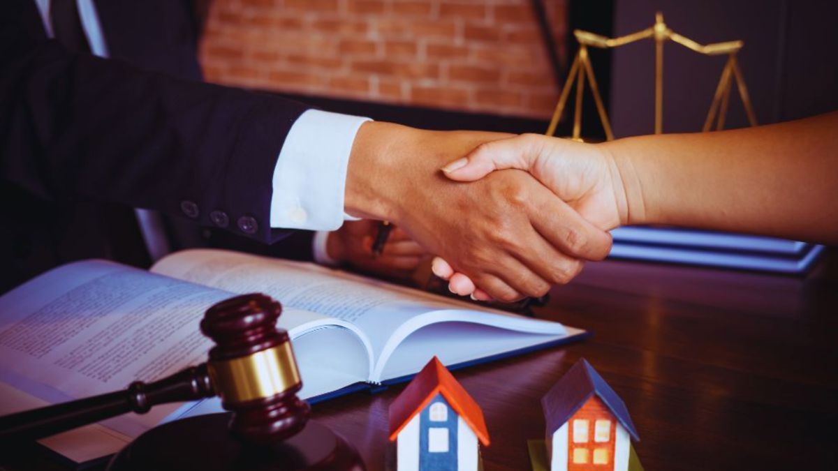 A lawyer and client shake hands across a desk with documents, a model house, a gavel, and scales of justice, representing a real estate closing or agreement.