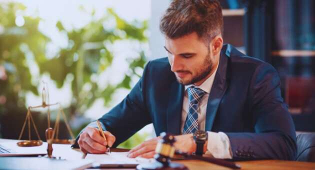A probate lawyer wearing a dark suit and tie is seated at a desk, writing on legal documents. A judge’s gavel, legal scale, and paperwork sit on the desk, suggesting a professional law office environment.