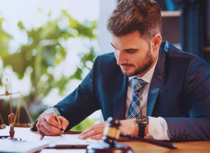 A probate lawyer wearing a dark suit and tie is seated at a desk, writing on legal documents. A judge’s gavel, legal scale, and paperwork sit on the desk, suggesting a professional law office environment.