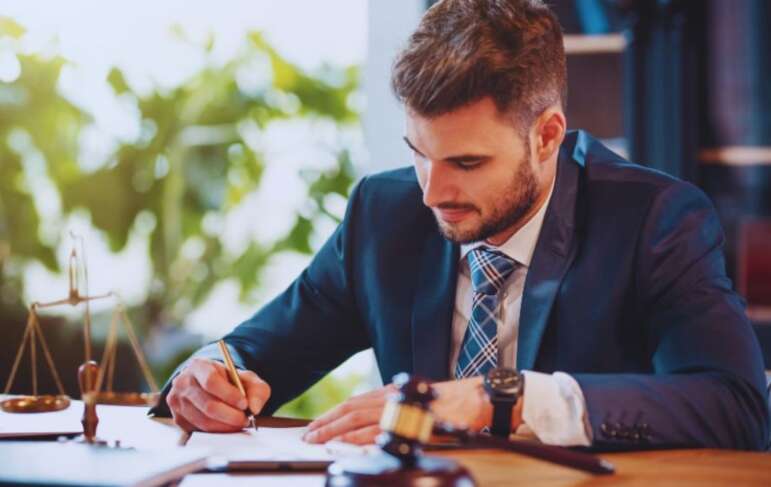 A probate lawyer wearing a dark suit and tie is seated at a desk, writing on legal documents. A judge’s gavel, legal scale, and paperwork sit on the desk, suggesting a professional law office environment.