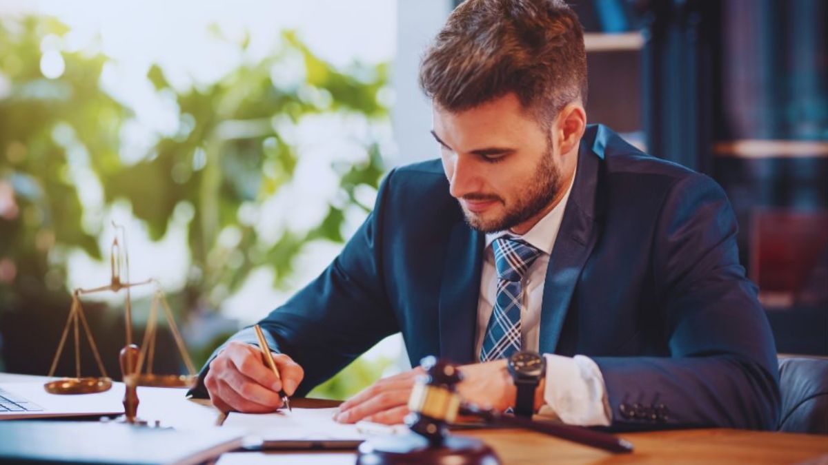 A probate lawyer wearing a dark suit and tie is seated at a desk, writing on legal documents. A judge’s gavel, legal scale, and paperwork sit on the desk, suggesting a professional law office environment.