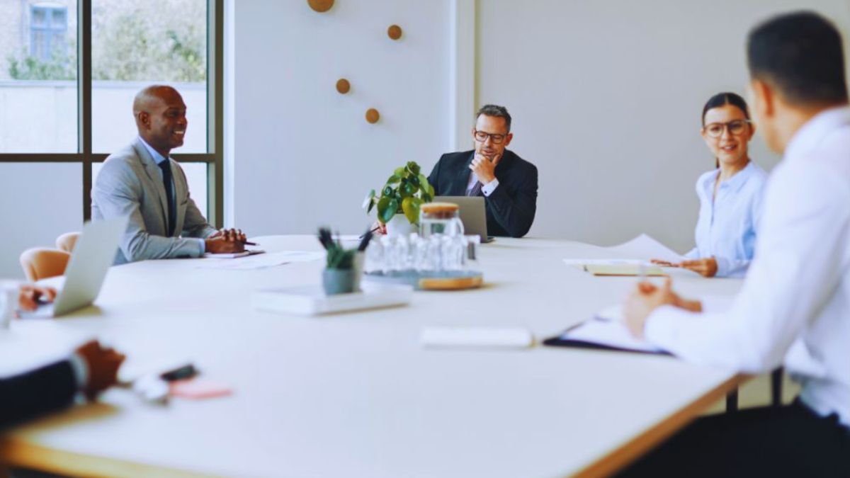 Business executives seated around a modern conference table discussing business formation strategy during a formal meeting, with laptops, documents, and glassware arranged in the center of the table.