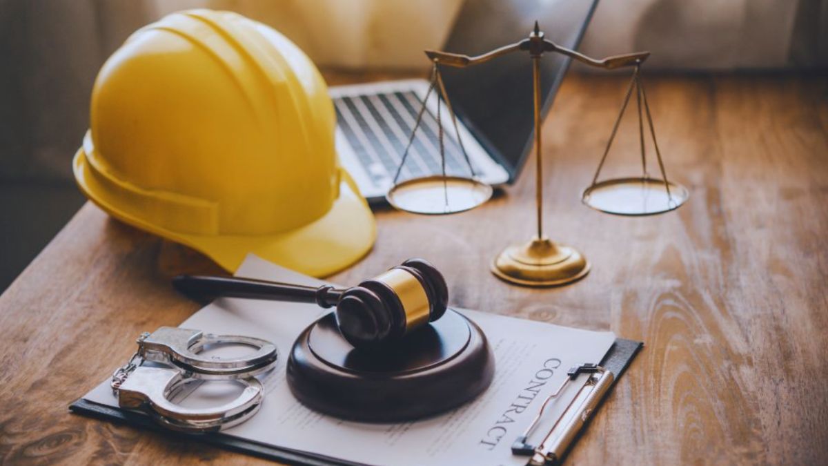 Yellow construction hard hat, gavel, handcuffs, and scales of justice on a wooden desk with a construction contract on a clipboard, representing legal help for construction contract disputes.