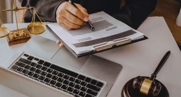 Debt collection lawyer in a suit reviewing and marking a legal document on a clipboard over a laptop, with scales of justice and a judge’s gavel on the desk.