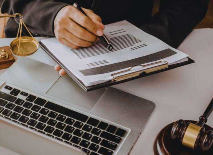 Debt collection lawyer in a suit reviewing and marking a legal document on a clipboard over a laptop, with scales of justice and a judge’s gavel on the desk.