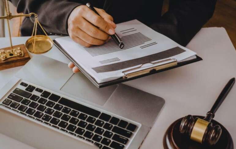 Debt collection lawyer in a suit reviewing and marking a legal document on a clipboard over a laptop, with scales of justice and a judge’s gavel on the desk.