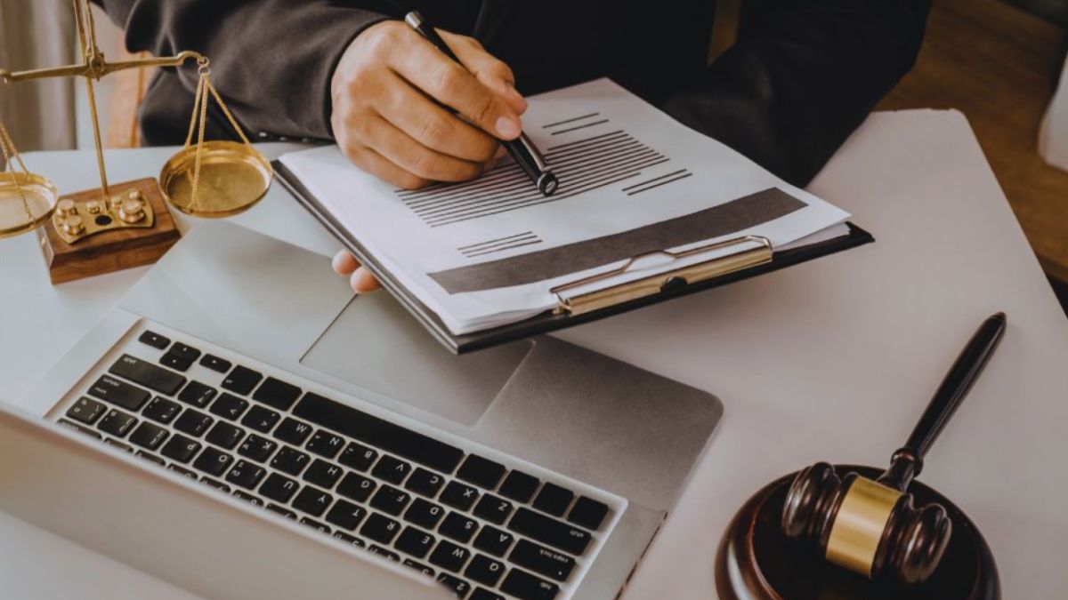 Debt collection lawyer in a suit reviewing and marking a legal document on a clipboard over a laptop, with scales of justice and a judge’s gavel on the desk.