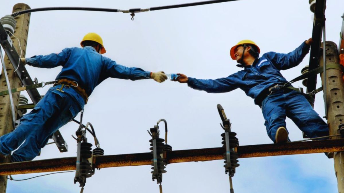 Two utility workers in safety harnesses and yellow helmets stand on high power lines, passing a tool between them while maintaining overhead electrical equipment, illustrating a utility easement in use.