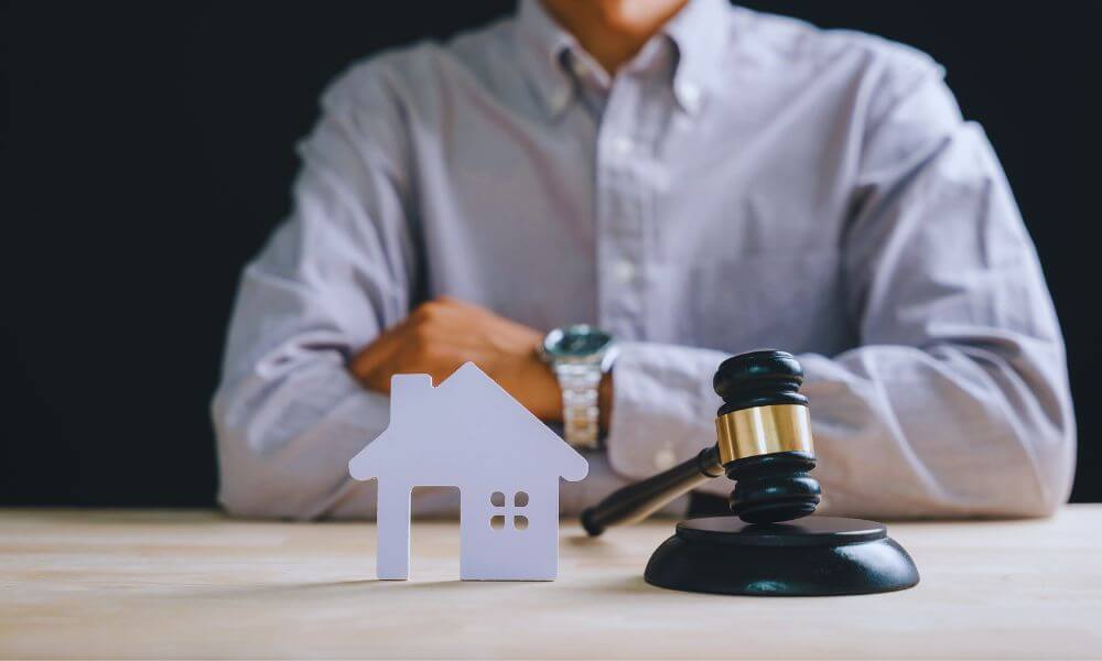 Attorney in a dress shirt sitting with arms crossed behind a small white house model and a judge’s gavel on a desk, representing property dispute legal services in Hilton Head Island, SC