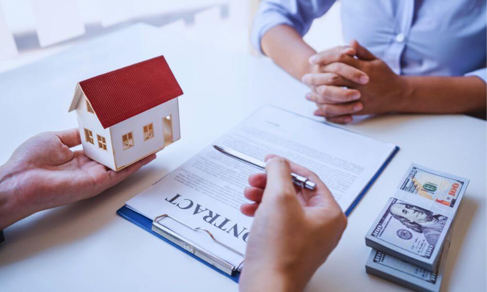 Home buyer and agent reviewing a real estate contract at a desk, with a small model house, pen, and stacks of cash symbolizing a home purchase.