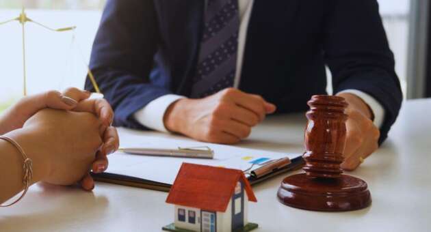 Real estate lawyer meeting with a client at a desk, with a small model house, legal documents, and a judge’s gavel in the foreground.