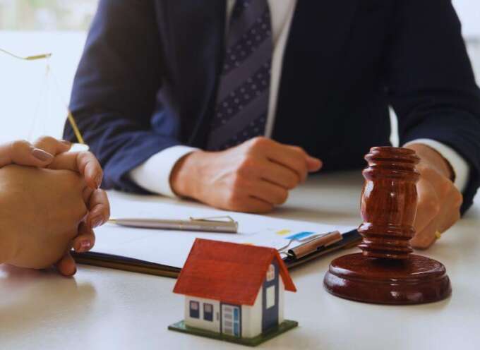 Real estate lawyer meeting with a client at a desk, with a small model house, legal documents, and a judge’s gavel in the foreground.