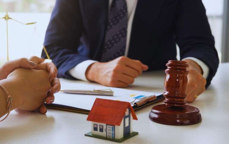 Real estate lawyer meeting with a client at a desk, with a small model house, legal documents, and a judge’s gavel in the foreground.