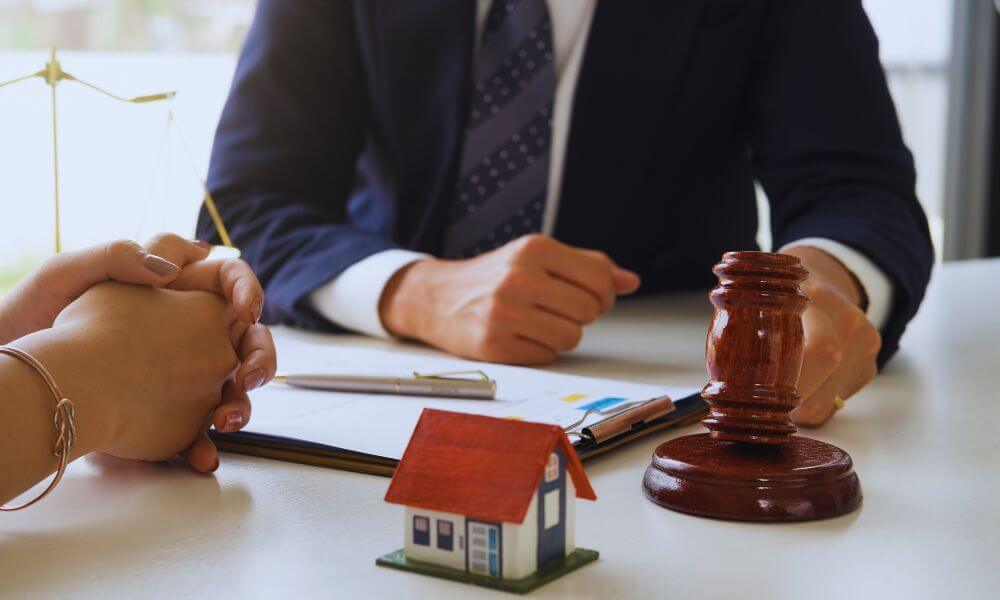 Real estate lawyer meeting with a client at a desk, with a small model house, legal documents, and a judge’s gavel in the foreground.