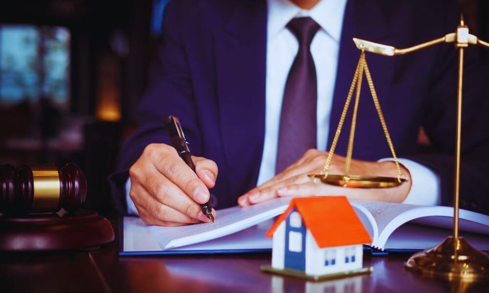 Hilton Head real estate attorney in a suit signing legal documents at a desk, with a model house, judge’s gavel, and golden scales of justice in the foreground.